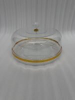 A clear acrylic cake dome with a gold-rimmed textured base and a crystal-style knob, showcasing a yellow sponge cake on a white marble kitchen counter.