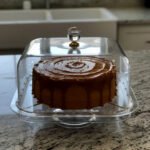 A versatile clear acrylic 2-in-1 serving dish shown in two uses: one as a divided tray filled with assorted dry fruits, nuts, and apricots, and the other as a pedestal cake stand holding a caramel-drizzled cake under a protective dome.