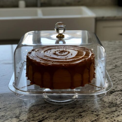 A versatile clear acrylic 2-in-1 serving dish shown in two uses: one as a divided tray filled with assorted dry fruits, nuts, and apricots, and the other as a pedestal cake stand holding a caramel-drizzled cake under a protective dome.