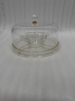 A versatile 2-in-1 clear acrylic serving dish shown in two settings: first as a pedestal cake stand holding a red velvet cake in a bright kitchen, and second as a flat sectional tray filled with almonds, pistachios, and dried apricots on a wooden coffee table.