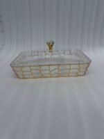 A 6-compartment clear acrylic dry fruit tray containing dates, apricots, almonds, pistachios, and raisins, held in a gold mesh frame on a dark wooden side table.