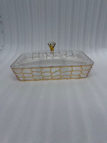 A 6-compartment clear acrylic dry fruit tray containing dates, apricots, almonds, pistachios, and raisins, held in a gold mesh frame on a dark wooden side table.