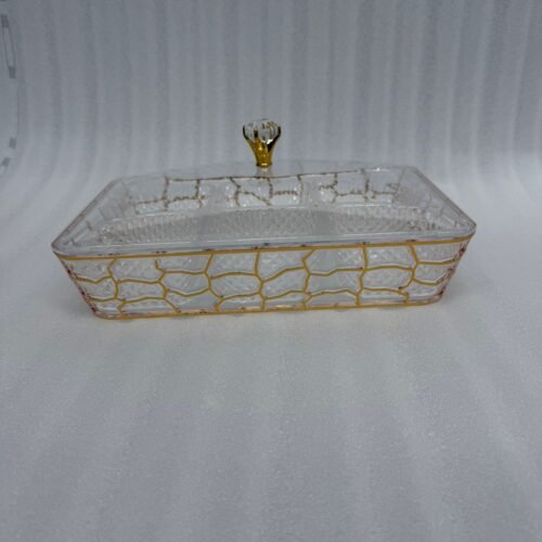 A 6-compartment clear acrylic dry fruit tray containing dates, apricots, almonds, pistachios, and raisins, held in a gold mesh frame on a dark wooden side table.