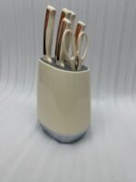 A 6-piece kitchen knife set featuring cream-colored ergonomic handles with rose gold accents, displayed both inside a matching compact storage block on a granite countertop and laid out on a white surface to show the different stainless steel blades and kitchen shears.