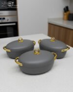 A set of three modern, oval-shaped insulated hotpots with gold handles and knobs, shown in chocolate brown, grey, and white colors on wooden and marble dining surfaces.