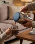 A 7-piece smoky grey glass water set featuring a large pitcher with a blue lid and six matching mugs with gold handles, shown on a rustic wooden dining table with steaming tea.