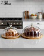 Two round cake stands of different sizes on a white kitchen counter, featuring natural wooden bamboo bases and clear ribbed acrylic dome covers with decorative crystal knobs, containing fruit cakes.