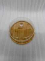 A white frosted layer cake displayed on a natural wooden bamboo base, covered by a clear circular acrylic dome with a round glass-like knob on a white marble kitchen counter.