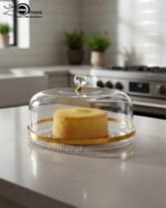 A clear acrylic cake dome with a gold-rimmed textured base and a crystal-style knob, showcasing a yellow sponge cake on a white marble kitchen counter.
