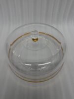 A clear acrylic cake dome with a gold-rimmed textured base and a crystal-style knob, showcasing a yellow sponge cake on a white marble kitchen counter.