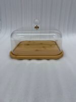 A chocolate cake with caramel drizzle sitting on a square bamboo wooden base, covered by a clear acrylic dome with a crystal handle on a white marble kitchen countertop.