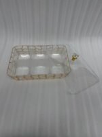 A 6-compartment clear acrylic dry fruit tray containing dates, apricots, almonds, pistachios, and raisins, held in a gold mesh frame on a dark wooden side table.