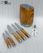 A 6-piece stainless steel kitchen knife set with light brown wood-grain handles and white kitchen shears, displayed both inside and next to a matching oval wood-patterned storage block on a granite countertop.