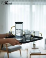 A person carrying a black serving tray with a modern smoke-grey glass water pitcher and six matching mugs with amber handles, filled with ice water in a bright, minimalist living room.