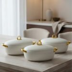 A set of three modern, oval-shaped insulated hotpots with gold handles and knobs, shown in chocolate brown, grey, and white colors on wooden and marble dining surfaces.