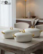 A set of three modern, oval-shaped insulated hotpots with gold handles and knobs, shown in chocolate brown, grey, and white colors on wooden and marble dining surfaces.
