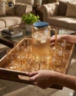 A 7-piece amber-tinted glass water set featuring a tall pitcher with a navy blue lid and six matching mugs arranged on a wooden table and a serving tray in a sunlit living room.
