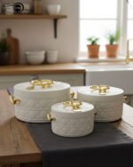 A set of three round, quilted insulated hotpots in varying sizes (1L, 2.5L, and 4L) with gold handles and lid accents, displayed on a kitchen counter and wooden dining table.