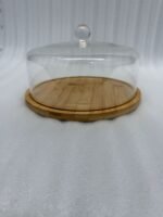 A white frosted layer cake displayed on a natural wooden bamboo base, covered by a clear circular acrylic dome with a round glass-like knob on a white marble kitchen counter.