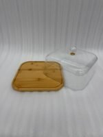 A chocolate cake with caramel drizzle sitting on a square bamboo wooden base, covered by a clear acrylic dome with a crystal handle on a white marble kitchen countertop.
