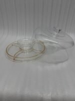 A versatile 2-in-1 clear acrylic serving set shown in two views: one as a divided platter filled with dates, raisins, and walnuts under a dome lid, and the other as a pedestal stand holding a layered chocolate cake on a marble kitchen counter.