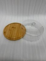 A white frosted layer cake displayed on a natural wooden bamboo base, covered by a clear circular acrylic dome with a round glass-like knob on a white marble kitchen counter.
