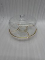 A versatile 2-in-1 clear acrylic serving set shown in two views: one as a divided platter filled with dates, raisins, and walnuts under a dome lid, and the other as a pedestal stand holding a layered chocolate cake on a marble kitchen counter.