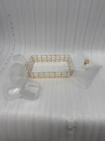 A 6-compartment clear acrylic dry fruit tray containing dates, apricots, almonds, pistachios, and raisins, held in a gold mesh frame on a dark wooden side table.