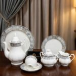 A luxury 24-piece white porcelain tea set featuring a grey marble effect border and intricate silver filigree patterns, displayed on a wooden surface against elegant neutral curtains.