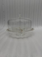 A versatile 2-in-1 clear acrylic serving dish shown in two settings: first as a pedestal cake stand holding a red velvet cake in a bright kitchen, and second as a flat sectional tray filled with almonds, pistachios, and dried apricots on a wooden coffee table.