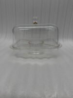 A versatile clear acrylic 2-in-1 serving dish shown in two uses: one as a divided tray filled with assorted dry fruits, nuts, and apricots, and the other as a pedestal cake stand holding a caramel-drizzled cake under a protective dome.