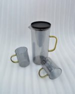 A person carrying a black serving tray with a modern smoke-grey glass water pitcher and six matching mugs with amber handles, filled with ice water in a bright, minimalist living room.