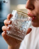 Close-up of a hand holding an RCR-inspired crystal water glass with a geometric starburst pattern and a decorative gold rim.