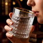 Close-up of a hand holding a diamond-textured crystal glass with a gold rim, filled with water, against a warm library background.