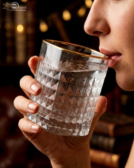 Close-up of a hand holding a diamond-textured crystal glass with a gold rim, filled with water, against a warm library background.