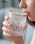Close-up of a person drinking from an RCR-inspired crystal water glass featuring a textured diamond-cut pattern and a luxurious golden rim.