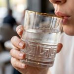 Close-up of a person drinking from a Versace-inspired crystal glass featuring an embossed Greek Key pattern and a luxury golden rim.