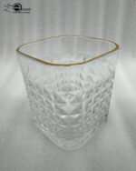 Close-up of a hand holding a diamond-textured crystal glass with a gold rim, filled with water, against a warm library background.