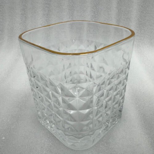 Close-up of a hand holding a diamond-textured crystal glass with a gold rim, filled with water, against a warm library background.