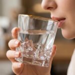 A close-up of a premium RCR-inspired crystal whiskey glass with a textured ice-like geometric base and a thin golden outline on the rim, being used to drink water.