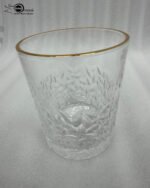 A close-up of a person holding a clear crystal water glass with a textured diamond pattern and a thin golden rim, showcasing the elegant light refraction through the water.