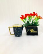 A luxury hexagonal ceramic coffee mug with gold marble veining, featuring a gold-plated square handle and rim, displayed next to a small potted plant with red flowers.
