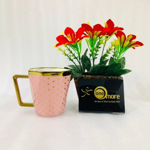 A modern hexagonal ceramic mug with gold polka dots and a gold handle, shown in two color options: blush pink and bright yellow, placed next to a small pot of red flowers.