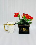 A luxury hexagonal ceramic coffee mug with gold marble veining, featuring a gold-plated square handle and rim, displayed next to a small potted plant with red flowers.