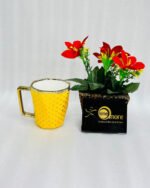 A modern hexagonal ceramic mug with gold polka dots and a gold handle, shown in two color options: blush pink and bright yellow, placed next to a small pot of red flowers.