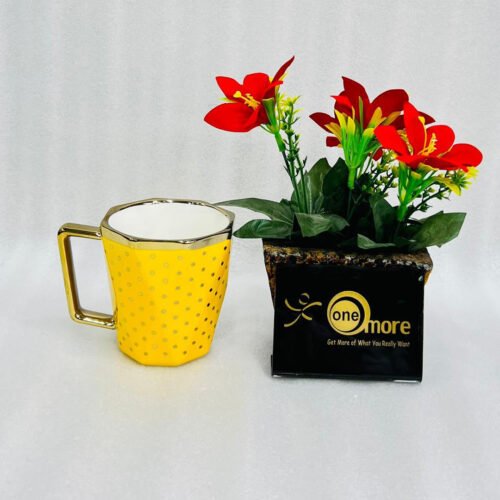 A modern hexagonal ceramic mug with gold polka dots and a gold handle, shown in two color options: blush pink and bright yellow, placed next to a small pot of red flowers.