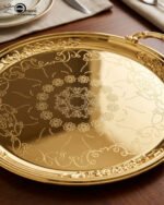A luxury set of three circular 18K gold-plated stainless steel serving trays featuring intricate floral engravings and ornate handles, displayed on a white runner over a wooden table.