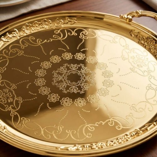 A luxury set of three circular 18K gold-plated stainless steel serving trays featuring intricate floral engravings and ornate handles, displayed on a white runner over a wooden table.