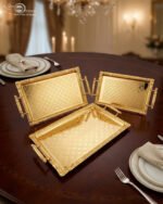 A luxury three-piece set of rectangular gold-plated stainless steel serving trays with intricate floral embossing and ornate vintage handles, displayed on a wooden dining table with elegant place settings.