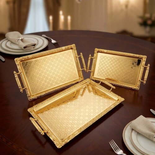 A luxury three-piece set of rectangular gold-plated stainless steel serving trays with intricate floral embossing and ornate vintage handles, displayed on a wooden dining table with elegant place settings.